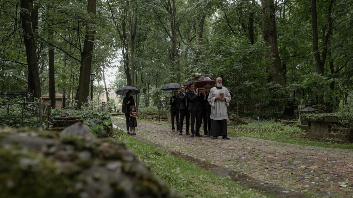 Mourners walk through a peaceful, wooded cemetery during a funeral procession.