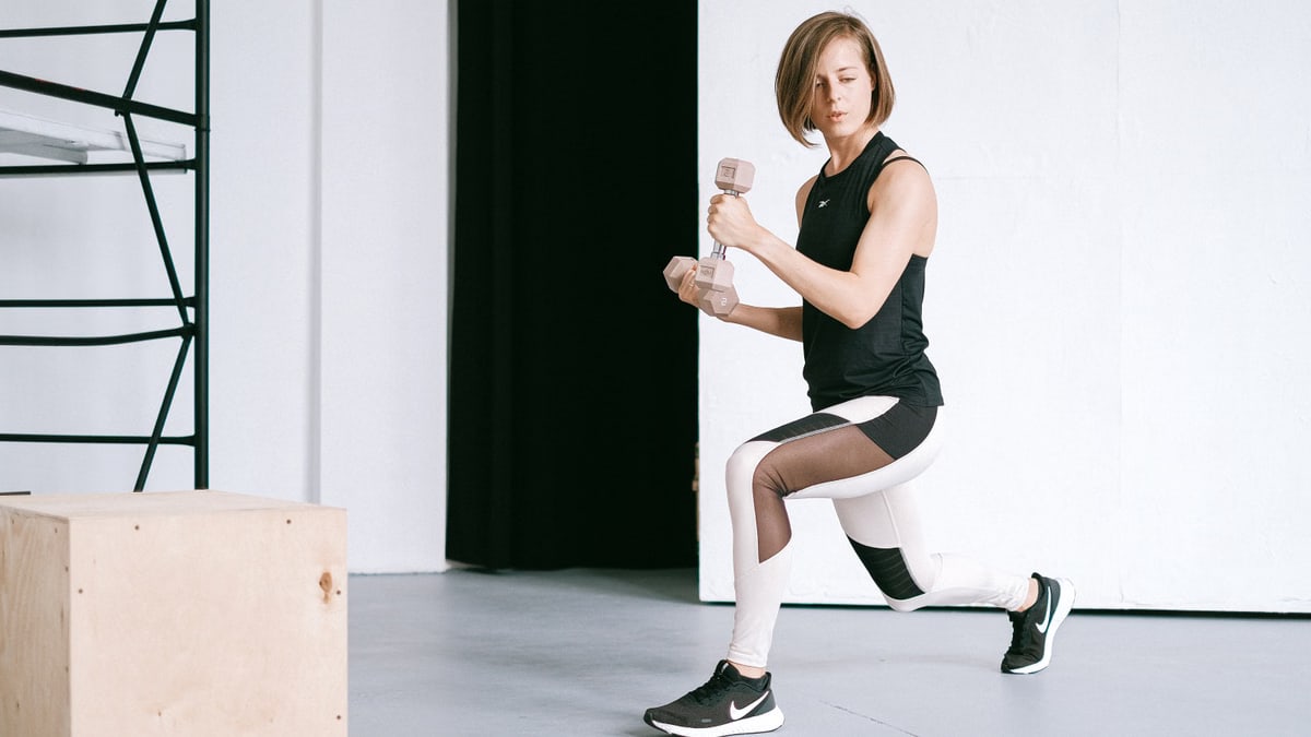 A young woman performing lunges with dumbbells in a modern indoor gym setting.